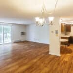 Open, unfurnished apartment with wood floors, a chandelier, sliding glass doors leading to a balcony, white walls, and an adjacent kitchen with dark cabinets and stainless steel appliances.