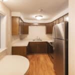 A small kitchen with wood cabinets, stainless steel appliances, light countertops, wooden floor, open shelving on the right, and a pass-through window to the left. The space is well-lit with overhead lighting.