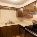 A small kitchen with brown wooden cabinets, beige countertops, a stainless steel stove and range hood, a dishwasher, and a single-basin sink under overhead cabinets. The walls are covered with light-patterned wallpaper.