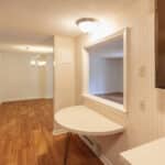 Kitchen with light wood flooring, a stainless steel refrigerator on the left, a pass-through window with a foldable table, and a view into a dining area with chandeliers and light-colored walls.
