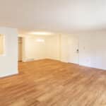 A spacious, empty room with light-colored walls and wood flooring. There is a small open window to a kitchen area, a white door, a chandelier, and a glimpse of a refrigerator through the window.