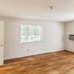 Empty room with light-colored walls, wood-look flooring, two windows with blinds, a wall-mounted air conditioner, baseboard heating, and an open door leading to a bathroom with tile flooring.