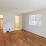 An empty room with light wood flooring, white walls, a large window with blinds, and two open doors leading to a walk-in closet and a bathroom with a vanity and mirror.
