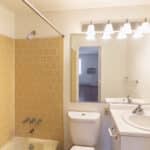 A bathroom with yellow tiled walls and a bathtub, a white toilet, a white vanity with a sink, a large mirror, and four-light vanity fixture above the mirror. The room appears clean and well-lit.