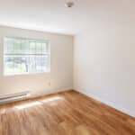 An empty room with light-colored walls, a large window letting in natural light, wood flooring, and a baseboard heater beneath the window. The room appears clean and unfurnished.