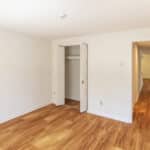 A bright, empty room with light-colored walls, wood flooring, an open closet with double doors, and a hallway leading to other rooms.