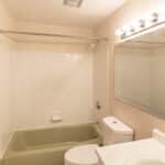 A small bathroom with beige wallpaper, a green bathtub-shower combo, a white toilet, a white sink, a large mirror, and bright vanity lights above the mirror.