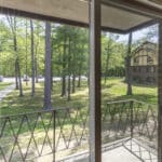 View through a glass sliding door showing a small balcony with black railing, overlooking a grassy area with trees and a sidewalk, and a brown and tan building in the background.