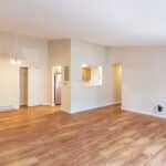 Spacious, empty room with light wood flooring, beige walls, a ceiling fan, open doorway to a kitchen, and natural light entering from the right. Kitchen features white appliances and cabinetry.