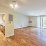 Spacious, empty living room with wood flooring, cream-colored walls, and sliding glass doors leading to a balcony. An open doorway shows a small kitchen with wooden cabinets. Ceiling lights are on.