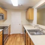 A modern kitchen with light wood cabinets, stainless steel appliances, a double sink, electric stovetop, microwave, and a white door at the back. The countertops are beige, and the floor is light wood.