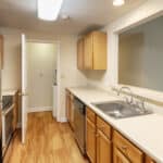 A galley kitchen with wood floors, light wood cabinets, a stainless steel refrigerator and oven, a dishwasher, double sink, and cream-colored countertops. A door is open at the far end, revealing a laundry area.