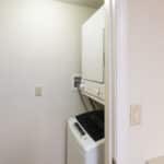 A small laundry room with a stacked washer and dryer, white walls, wire shelves on the left, and a light switch on the right wall near wooden cabinets and a countertop.