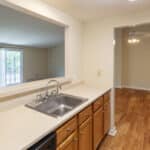 A kitchen with light wood cabinets, a stainless steel sink, and tan countertops. An open window connects to the living area with large windows. The kitchen leads to a dining area with wood flooring and a ceiling fan.