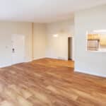 An empty apartment interior with light wood flooring, beige walls, a white door, and an open kitchen featuring stainless steel appliances and light wood cabinets visible through a large pass-through window.