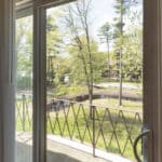A glass sliding door opens to a small balcony with a metal railing, overlooking a grassy area with trees and a curved pathway outside on a sunny day.