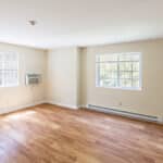 A bright, empty room with light beige walls, wood flooring, two large windows, a wall-mounted air conditioner, baseboard heater, and an open white door. Sunlight streams through the windows.