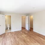 Bright empty bedroom with light wood flooring, cream-colored walls, large window, open closet, and en suite bathroom with the door open showing a toilet.
