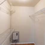 Empty walk-in closet with white wire shelving on both sides, beige walls, a light wood floor, and an electric panel on the left wall.