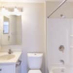 A clean, bright bathroom with a white sink and cabinet, a toilet, a large mirror, overhead lights, and a white bathtub with a shower curtain rod and built-in shelves against beige walls.
