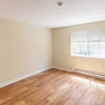 An empty room with beige walls, a wooden floor, a white baseboard heater, and a window with partially open blinds letting in sunlight.