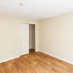 Empty room with light beige walls, a small closet with sliding door, and wood-style flooring. An open doorway leads to a hallway with similar flooring and another white door visible in the background.