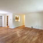 Empty living room with light-colored walls, wood flooring, a small open kitchen window, a closet with white doors, and a ceiling light fixture. A few electrical cables are visible against one wall.