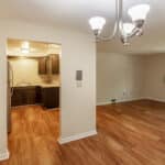 A bright, empty room with hardwood floors, beige walls, a ceiling light fixture, and an open doorway leading to a small kitchen with dark cabinets and stainless steel appliances. A window lets in natural light.