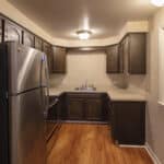 A small kitchen with dark wooden cabinets, stainless steel refrigerator, light countertops, wood flooring, and overhead lighting. There is a serving window on the right and a sink under the cabinets at the back.