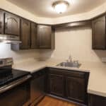 A kitchen with dark wooden cabinets, beige countertops, stainless steel appliances, a built-in stove, a dishwasher, a double sink, and overhead lighting. The walls are light-colored with a textured finish.