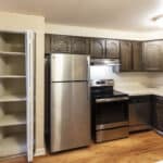 A kitchen with stainless steel appliances, dark wooden cabinets, a countertop with a sink, an open pantry with shelves, and wood-style flooring. Ceiling light fixture provides illumination.