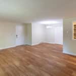 An empty living room with light-colored walls and wood flooring. There is a white entry door, a closet with double doors, a small chandelier, and a serving window opening to a kitchen with stainless steel appliances.