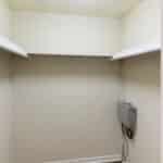 Empty walk-in closet with cream-colored walls, white shelves on both sides, a slatted door partially open, and an electrical box mounted on the lower right wall. The space is well-lit and clean.