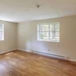 A bright, empty room with light-colored walls, two windows with blinds, wood flooring, a wall-mounted air conditioner, baseboard heater, and an open white door. Sunlight streams in through the windows.