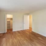 Empty room with light-colored walls, wood flooring, a large window on the left, an open closet with shelves, and an open door leading to another room. Natural light fills the space.