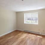 An empty room with light-colored walls, wood-style flooring, a white baseboard heater, and a window with blinds letting in sunlight. The room is clean and uncluttered, with natural light on the floor.