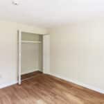 Empty room with light-colored walls, wood flooring, an open closet with white folding doors, and a doorway leading to a hallway with similar flooring. The room is well-lit with natural light.