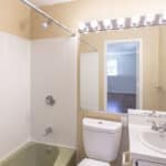 A small bathroom with a green bathtub, white toilet, and white sink. A large mirror above the sink reflects a lit vanity light bar and an adjacent room with wooden floors and a window.