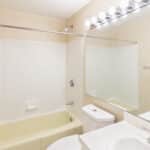 A clean, beige bathroom with a bathtub and shower combo, a white toilet, a sink with a countertop, a large wall mirror, and a row of lights above the mirror. The walls and tub are a light, neutral color.