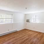Empty room with white walls, wood-style flooring, two windows with blinds, a wall-mounted air conditioner, and a white door slightly open on the left side. Natural light brightens the space.