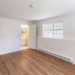 A bright, empty room with white walls, wood flooring, a window with blinds, two doors—one leading to a bathroom with a vanity and mirror, and a baseboard heater along the wall.