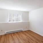 Empty room with white walls, a textured ceiling, wood-style flooring, a single window letting in natural light, and a baseboard heater below the window.