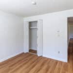 Empty room with white walls, light wood flooring, open double-door closet, and an open door leading to a hallway. The space is bright and clean, with simple, modern finishes.