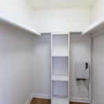 A small, empty walk-in closet with white walls, built-in upper shelves on both sides, a vertical shelving unit in the center, and a utility box with exposed wires on the right wall.