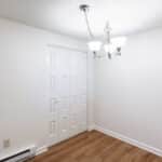 A small, empty room with white walls, a wooden floor, a double-door closet, and a modern ceiling light fixture with three frosted glass shades.