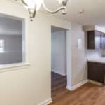 A small kitchen with dark cabinets and light countertops is visible next to a dining area with a decorative light fixture. A window opens to another room with large windows, and wood flooring runs throughout the space.