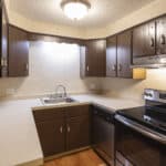 Small kitchen with brown cabinets, beige countertops, a stainless steel sink, black stove, dishwasher, and refrigerator, under a ceiling light fixture. The walls are light-colored with a subtle pattern.