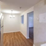A small kitchen with wood flooring, stainless steel refrigerator, beige walls, a white countertop, and a dining area with a chandelier. An open window leads to a bright living room with wood floors.