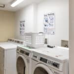 A small laundry room with several white washing machines and dryers, a white laundry basket on top, instructional posters on the wall, and folded laundry items nearby. The room has tiled flooring and bright lighting.