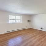 Empty room with light-colored walls, a large window, wood laminate flooring, baseboard heaters, and a wall-mounted air conditioning unit. Some outlets and cords are visible on the walls.